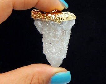 May include: A white quartz crystal pendant with a gold-plated top. The crystal is a cluster of small crystals that form a point. The pendant is held in a hand with blue nail polish.