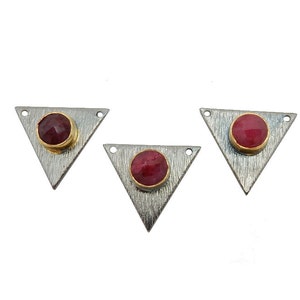 May include: Three silver-tone triangle pendants with a textured finish. Each pendant features a round, deep red gemstone set in a gold-tone bezel. Small holes are drilled at the top corners.