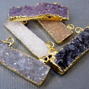 Könnte beinhalten: Eine Ansammlung von fünf rechteckigen Stabanhängern mit texturierten, kristallinen Oberflächen in den Farben Violett, Weiß, Pfirsich und Dunkelgrau. Jeder Anhänger ist mit einem goldfarbenen Metallrand eingefasst und hat eine kleine Öse.
