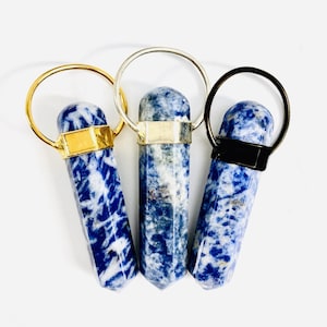 Puede incluir: Tres colgantes de piedra azul y blanca con anillos de metal dorado, plateado y negro. Los colgantes tienen todos la forma de un rectángulo largo y delgado con un extremo puntiagudo.