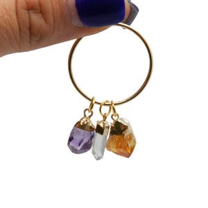 May include: Gold hoop earrings with three dangling charms: a purple amethyst, a clear quartz crystal, and a yellow citrine.
