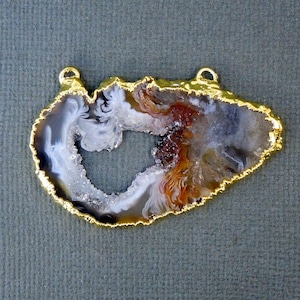 Puede incluir: Un colgante de &aacute;gata con borde dorado, que presenta una rebanada de &aacute;gata natural de color blanco, marr&oacute;n y gris. La &aacute;gata tiene un gran centro abierto en forma de coraz&oacute;n.