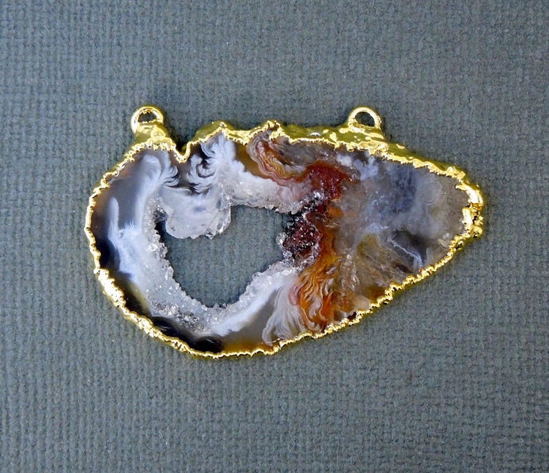 Puede incluir: Un colgante de &aacute;gata con borde dorado, que presenta una rebanada de &aacute;gata natural de color blanco, marr&oacute;n y gris. La &aacute;gata tiene un gran centro abierto en forma de coraz&oacute;n.