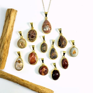 May include: A collection of 12 gold-toned pendants featuring various shades of brown, red, and white opal stones. The pendants are arranged on a white surface with a wooden stick in the background.