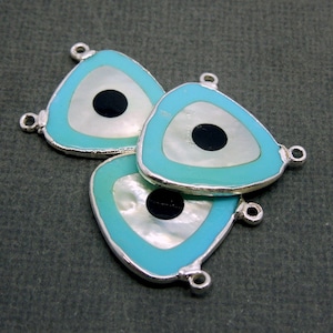 May include: Three turquoise blue triangle-shaped charms with a white and black evil eye design. Each charm has two silver jump rings.