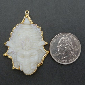 Op de afbeelding: Witte gegraveerde hanger met gouden rand, die een godheid met een olifantenkop voorstelt. De hanger is ongeveer zo groot als een Nederlandse 2 euro munt.
