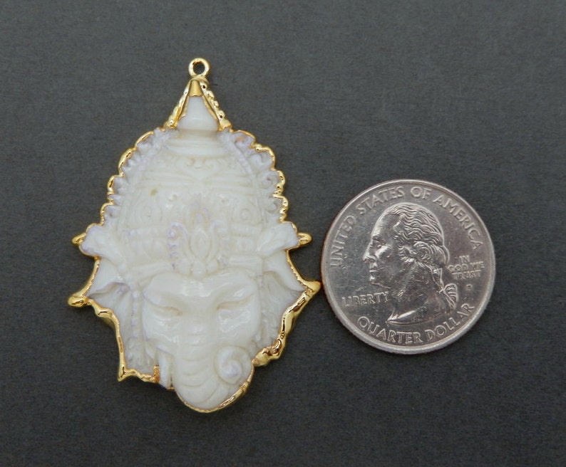 Op de afbeelding: Witte gegraveerde hanger met gouden rand, die een godheid met een olifantenkop voorstelt. De hanger is ongeveer zo groot als een Nederlandse 2 euro munt.