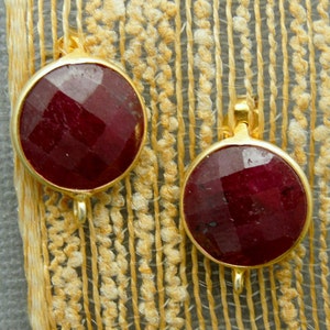 Op de afbeelding: Twee ronde rode edelsteen hangers met goudkleurige metalen instellingen en lussen. De edelstenen zijn gefacetteerd en hebben een glanzende afwerking.