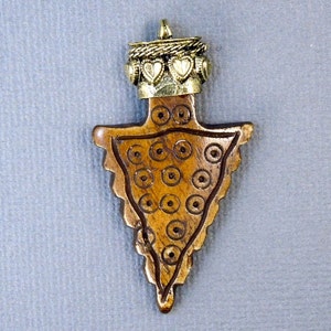 Op de afbeelding: Een houten pijlpunt hanger met een goudkleurig metalen bovenkant met een decoratief ontwerp met harten. De pijlpunt is gegraveerd met een herhalend cirkelpatroon.