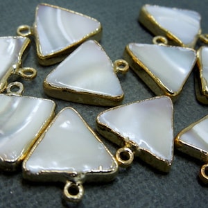 Puede incluir: Primer plano de varios colgantes triangulares con una superficie blanca nacarada y un borde dorado. Cada colgante tiene un pequeño lazo en la parte superior para sujetarlo a una cadena u otro componente de joyería. Los colgantes son probablemente para la fabricación de joyas.