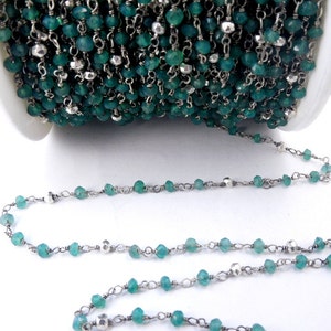 May include: A green gemstone and silver chain, perfect for making jewelry. The chain is made of small silver beads and green gemstones. The chain is coiled up on a white surface.