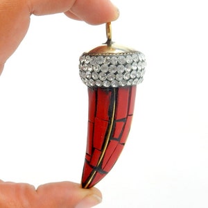 Op de afbeelding: Een rode en gouden hanger in de vorm van een tand, met een heldere kristallen bovenkant. De tand is gemaakt van rood koraal en heeft een gouden rand.