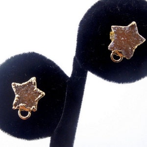 May include: Two gold star-shaped earring charms with a light brown, sparkly stone. The charms are on a black velvet display stand.