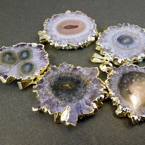 Puede incluir: Cinco colgantes de cristal de amatista con bordes chapados en oro. Cada colgante presenta un patrón único y natural de morado, blanco y marrón.