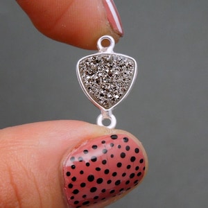 Puede incluir: Pendiente de plata con un dije de forma triangular que presenta una piedra gris oscura brillante. El dije está unido a un lazo de plata.
