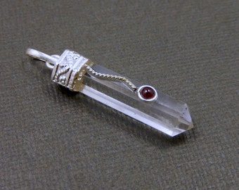 May include: A clear quartz crystal pendant with a silver setting and a small red gemstone accent. The pendant is on a grey background.