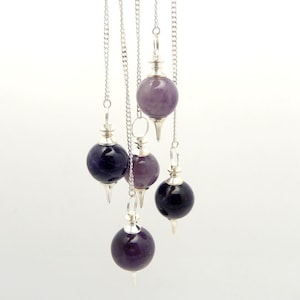 Puede incluir: Cinco péndulos de cristal de amatista y negro con cadenas plateadas que cuelgan de un fondo blanco.