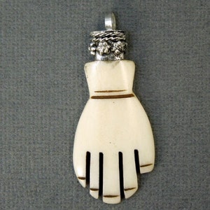 May include: A white bone hand pendant with a silver top. The hand has five fingers and a brown line around the wrist. The silver top has a rope design and small decorative balls.