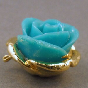 May include: A turquoise blue rose charm with a gold-tone setting and a loop for attaching to a necklace or bracelet.