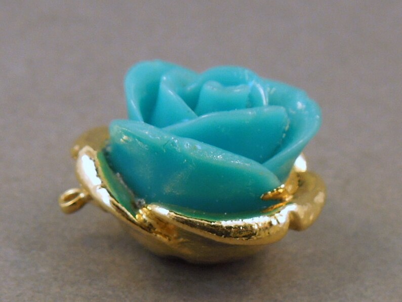 May include: A turquoise blue rose charm with a gold-tone setting and a loop for attaching to a necklace or bracelet.