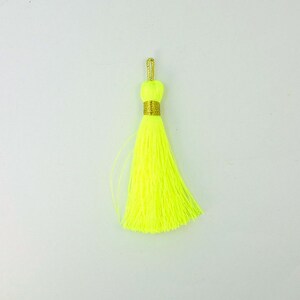 Puede incluir: Un borla de color amarillo neón brillante con un ribete dorado. La borla está hecha de hilo suave y esponjoso y tiene una larga franja fluida.