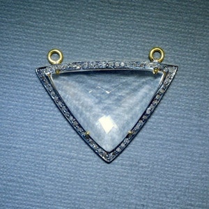 Puede incluir: Un colgante triangular con un centro de cristal transparente rodeado por un borde de pequeños diamantes. El colgante tiene dos bucles dorados en la parte superior para sujetar una cadena.