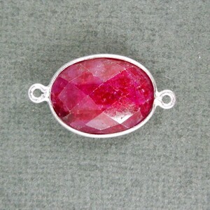 May include: A silver oval-shaped connector with a red ruby gemstone in the center. The connector has two loops on either side for attaching to jewelry.