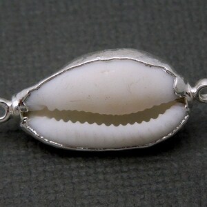 Könnte beinhalten: Eine weiße Kaurischnecke mit silbernen Akzenten und zwei Schlaufen zum Anbringen an einer Halskette oder einem Armband.