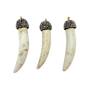 Op de afbeelding: Drie witte, tandvormige hangers met gouden toppen en zilveren strasssteentjes.