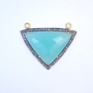 Puede incluir: Un colgante triangular con una piedra preciosa azul claro engastada en un marco de plata con un borde de diamantes. El colgante tiene dos bucles dorados en la parte superior para sujetar una cadena.