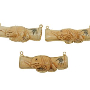 Op de afbeelding: Drie gouden hangers met gesneden bamboe-ontwerpen in ivoorwit. Elke hanger heeft een vis met zwarte details en een gouden lus om op te hangen.
