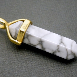 May include: A white howlite gemstone pendant with gold-toned metal accents. The pendant is shaped like a point and has a loop at the top for attaching to a chain.