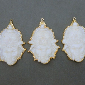 Op de afbeelding: Drie witte hangers met gouden versieringen, elk met een sierlijk, gedetailleerd ontwerp. Elke hanger heeft een gegraveerd gezicht met een kroon en ingewikkelde details.