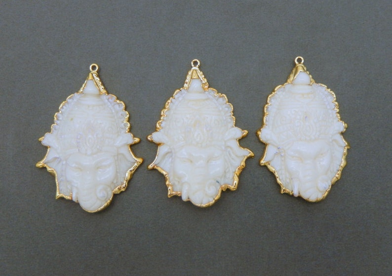 Op de afbeelding: Drie witte hangers met gouden versieringen, elk met een sierlijk, gedetailleerd ontwerp. Elke hanger heeft een gegraveerd gezicht met een kroon en ingewikkelde details.