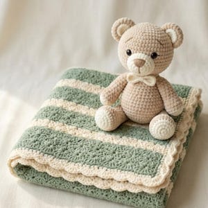 Può includere: Un orsacchiotto all'uncinetto color beige chiaro con un piccolo papillon seduto su una coperta piegata. La coperta presenta strisce orizzontali alternate verde salvia e crema, con un motivo a stella testurizzato e un bordo smerlato color crema.