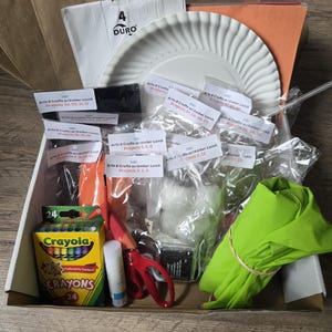 May include: A cardboard box containing various arts and crafts supplies, including a box of 24 Crayola crayons, a white paper plate, brown paper bags, a glue stick, red scissors, a lime green fabric piece, and multiple plastic bags with labels.