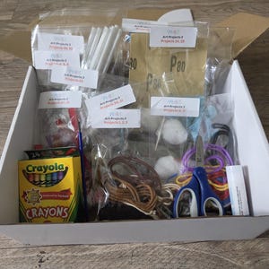 May include: A white cardboard box containing various art supplies in clear plastic bags, including a 24-count box of Crayola crayons, blue-handled scissors, a glue stick, sandpaper labeled P80, white sticks, yarn, and labels marked Art Projects 3.