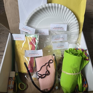 May include: A white cardboard box containing various art supplies including a white paper plate, a glue stick, green scissors, a roll of bright green fabric, felt pieces, black yarn, and several clear plastic bags labeled Art Projects K4.