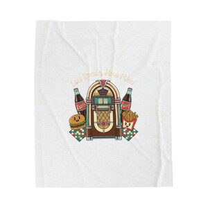 May include: A white blanket featuring a retro diner design. The design includes a jukebox, bottles of soda, a hamburger, and fries. The text "Less Drama, More Fries" is printed above the jukebox in gold lettering.