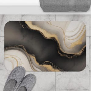 May include: A rectangular bath mat with a marbled design in black, gold, and white. The mat features flowing, abstract patterns with gold accents. Gray slippers are in the foreground, and a gray towel is in the background.