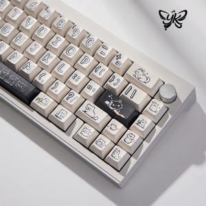 May include: A white mechanical keyboard with custom keycaps featuring black line art illustrations of cats. The keyboard has a silver knob and a black space bar. The keys are off-white with black lettering.