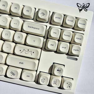 Könnte beinhalten: Nahaufnahme einer cremefarbenen Tastatur mit individuellen Tastenkappen. Die Tastenkappen zeigen schwarze Strichzeichnungen und Text, darunter "backspace", "shift" und Funktionstasten. Ein Schmetterlingslogo befindet sich oben rechts.