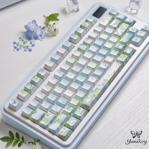 Può includere: Una tastiera meccanica azzurra e bianca con tasti floreali. I tasti presentano un design di fiori blu e verdi. La tastiera ha una cornice azzurra e un piccolo schermo. Il marchio "YuniKey" è visibile nell'angolo in basso a destra.