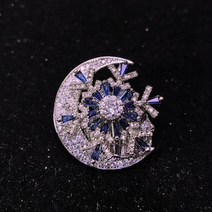 May include: A silver-toned brooch in the shape of a crescent moon and a snowflake. The brooch features a central round, clear stone surrounded by blue and clear stones. The crescent moon is encrusted with clear stones.