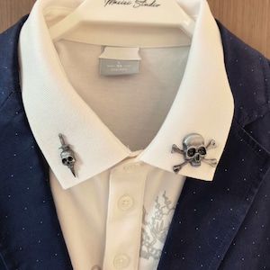 May include: A white collared shirt with two skull-themed collar pins, one with a dagger and the other with crossbones. The shirt is paired with a navy blue blazer with white polka dots. The hanger reads "Music Studio".