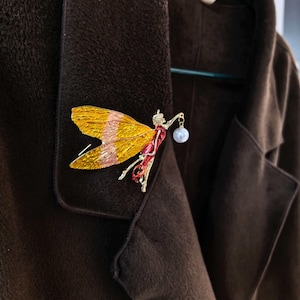 May include: A gold-toned dragonfly brooch with yellow and pink wings, red accents, and a pearl. The brooch is pinned to a brown coat. The dragonfly is holding a pearl on a chain.