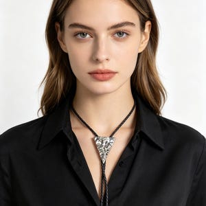 May include: A silver bolo tie with a triangular pendant featuring a detailed design. The tie has a black braided cord and silver tips. The person in the image is wearing a black button-down shirt.