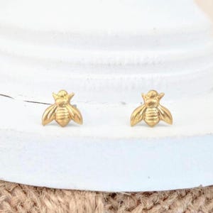 Könnte beinhalten: Ein Paar kleine Ohrstecker in Form einer goldenen Biene. Jeder Ohrstecker zeigt ein detailliertes, geprägtes Bienendesign mit ausgebreiteten Flügeln und einem segmentierten Körper, gefertigt mit einer glatten, polierten Oberfläche, die das Licht reflektiert.