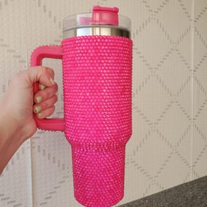 Pu&ograve; includere: Un grande bicchiere rosa acceso con un manico, ricoperto di piccoli abbellimenti scintillanti. Il bicchiere ha un coperchio trasparente con una chiusura a ribalta rosa. Anche il manico &egrave; rosa.