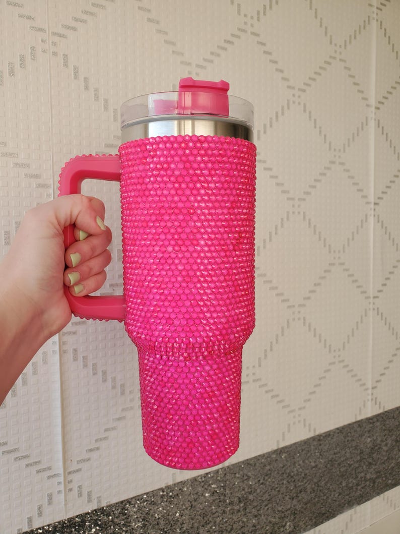 Pu&ograve; includere: Un grande bicchiere rosa acceso con un manico, ricoperto di piccoli abbellimenti scintillanti. Il bicchiere ha un coperchio trasparente con una chiusura a ribalta rosa. Anche il manico &egrave; rosa.